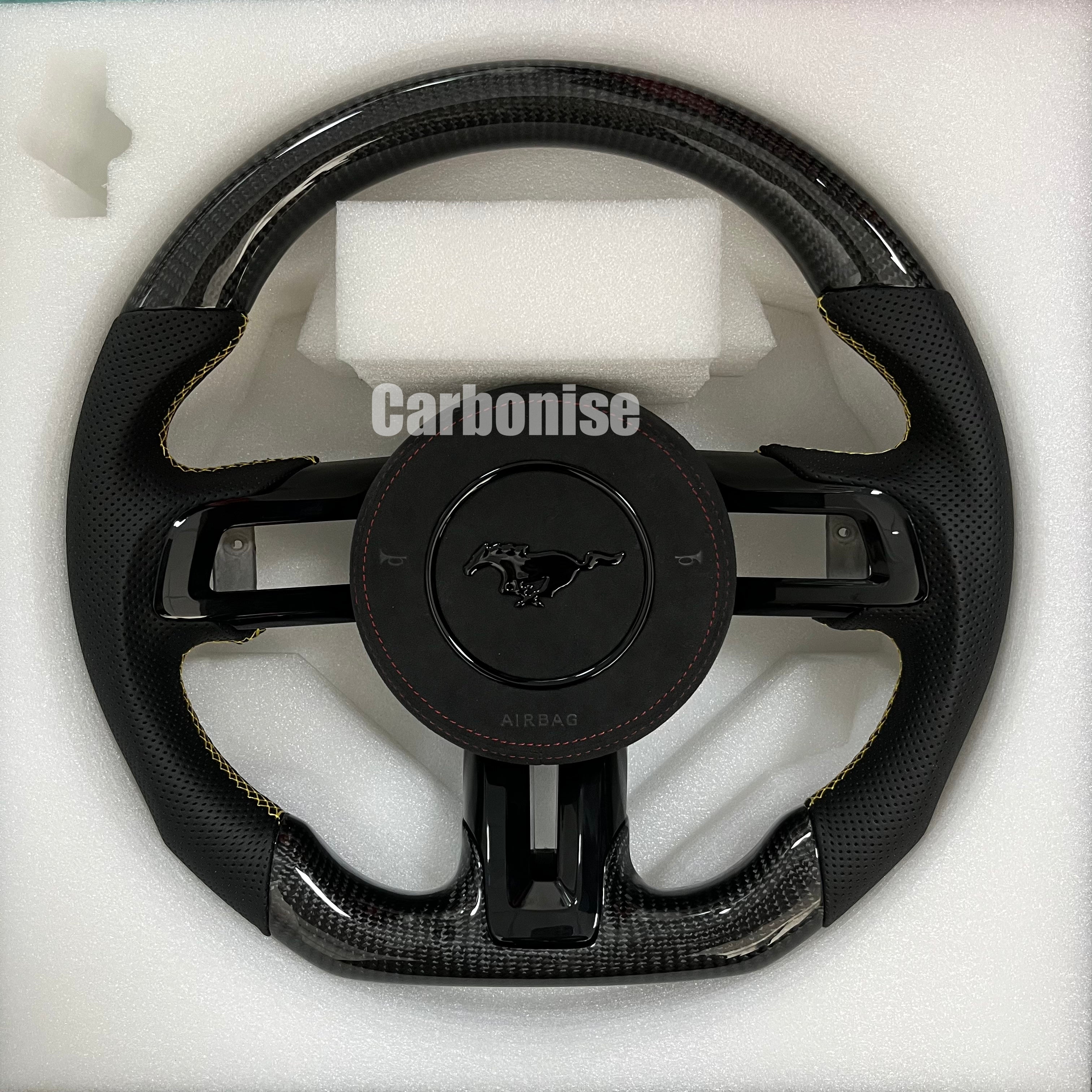 Ford Mustang carbon fibre steering wheel with perforated black leather grips, yellow stitching accents, round design, and OEM Mustang airbag cover.