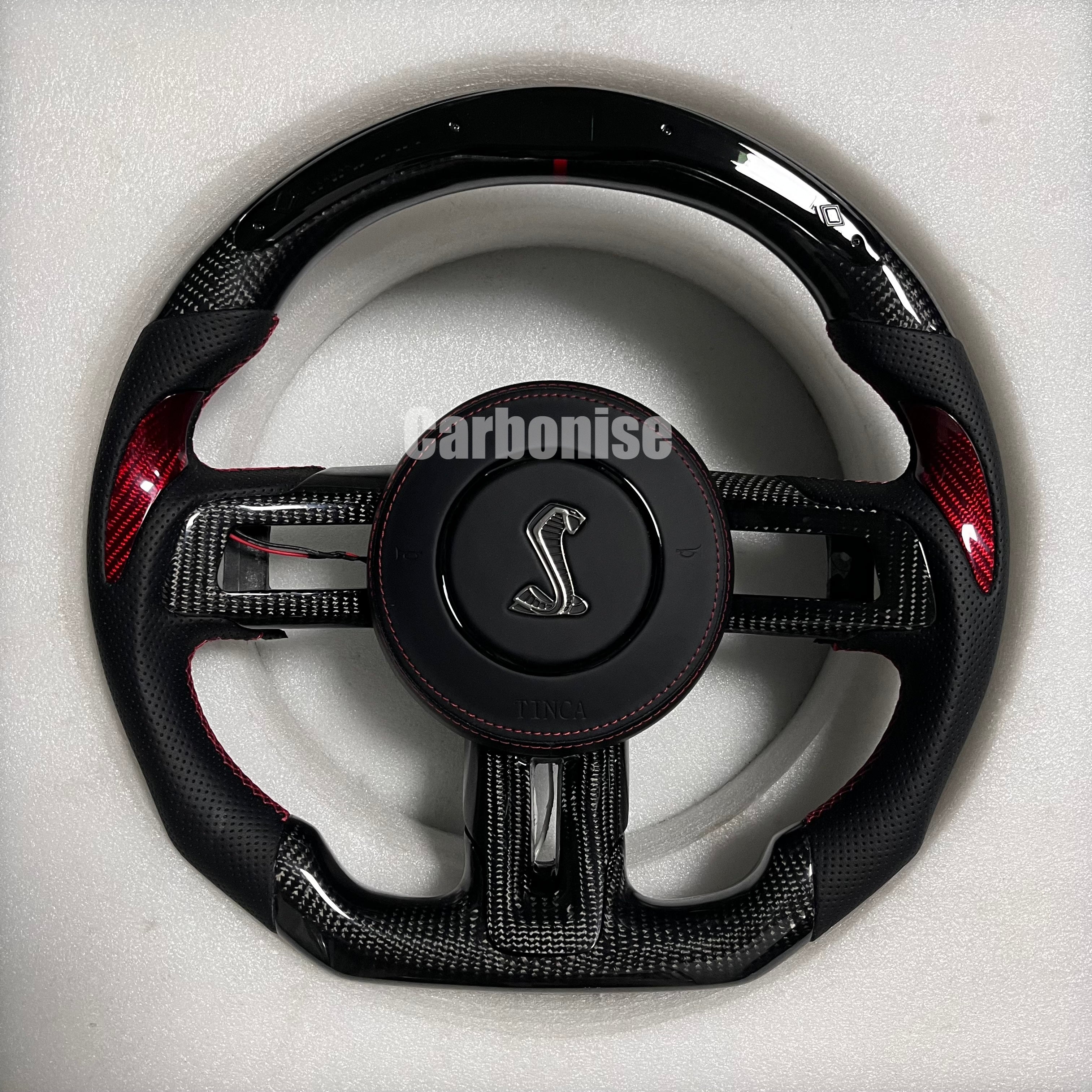 Ford Mustang Shelby carbon fibre steering wheel with red carbon accent inlays, perforated black leather grips, red stitching, gloss black carbon top and bottom, and Shelby Cobra airbag.