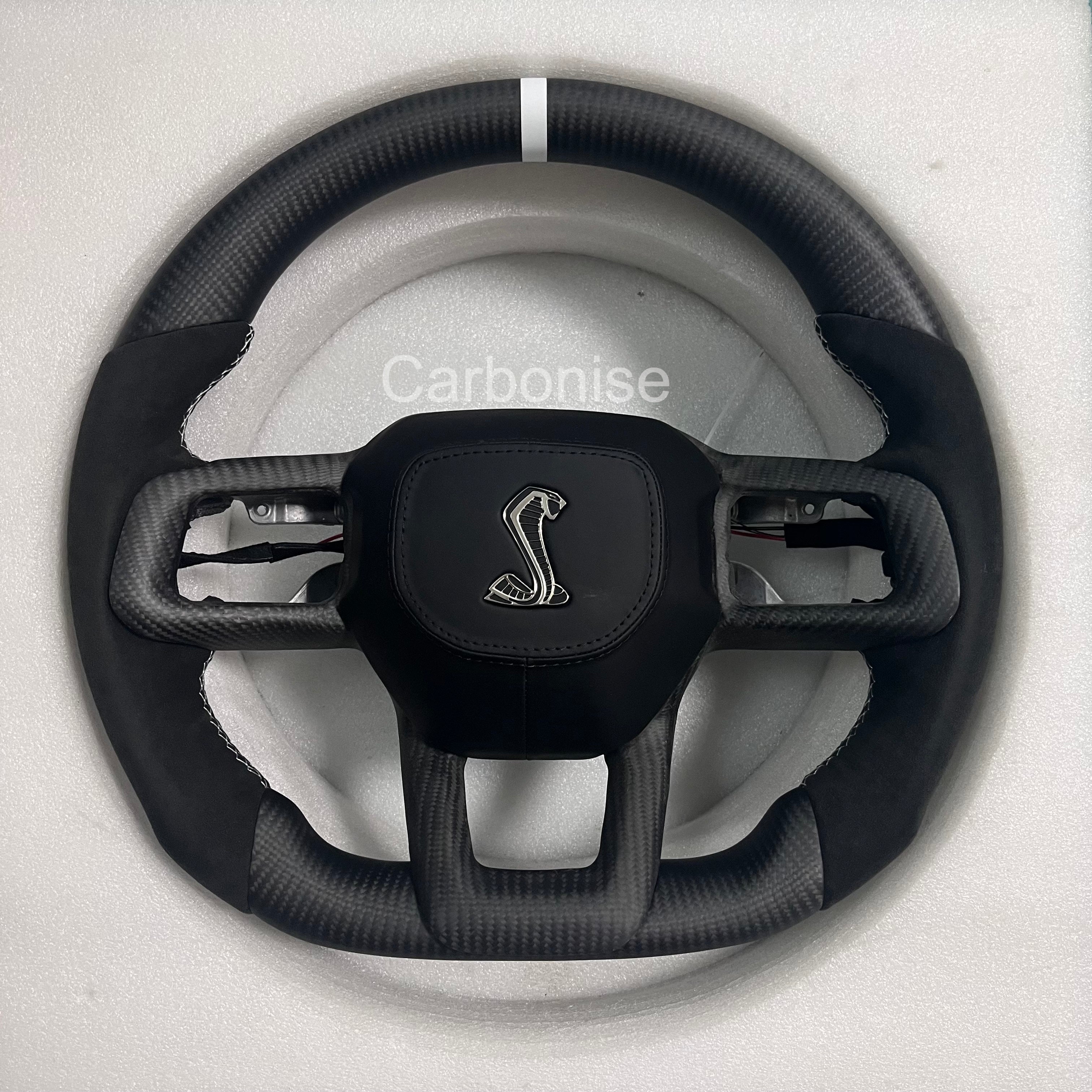 Ford Mustang Shelby matte carbon fibre steering wheel with Alcantara side grips, white centre stripe, round design, and Shelby Cobra airbag.