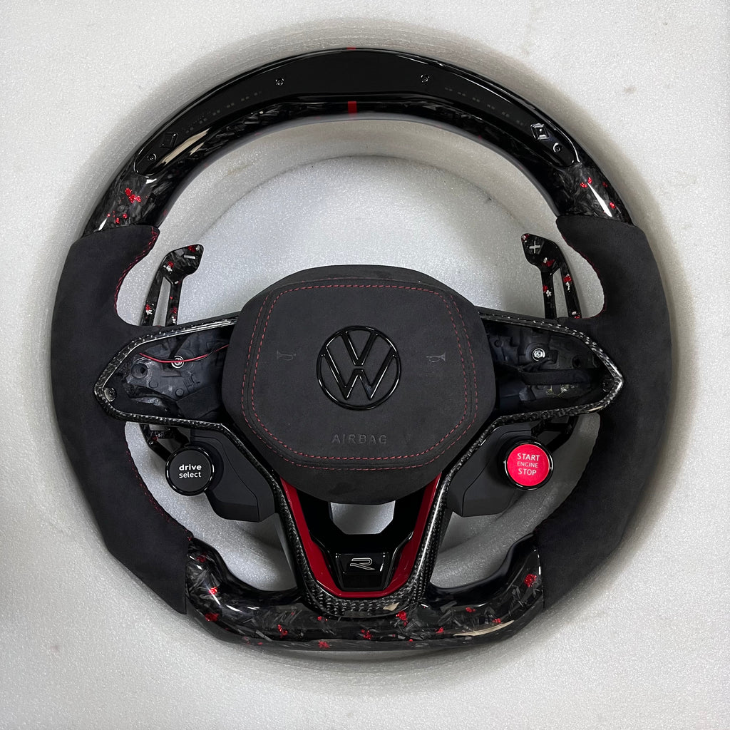 Custom Volkswagen performance steering wheel featuring forged carbon fiber with red flakes, integrated R8-style red ignition buttons, and black Alcantara side grips.